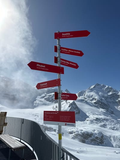 The image displays a metallic signpost featuring multiple red directional signs set against a snow-covered mountain landscape under a clear blue sky. This scene is located on a high-altitude observation deck in the Bernina Alps, near Pontresina, Switzerland.
The central subject is a grey metal pole with six red, arrow-shaped signs, each displaying a city name and an approximate flight duration in German ("Flugstunden"):
- "Seattle 10:30 Flugstunden" points upwards and right.
- "Halifax (Nova Scotia) 7:15 Flugstunden" points horizontally right.
- "Tampa Bay (Florida) 10:15 Flugstunden" points downwards and right.
- "Phuket 10:50 Flugstunden" points horizontally left.
- "Kapstadt 10:55 Flugstunden" points downwards left.
- "Punta Cana 9:50 Flugstunden" points downwards right, below Kapstadt.
Below these, a rectangular red information sign faces the viewer, detailing "Piz Bernina und Piz Palü" with their altitudes (4048m and 3900m respectively), a "Reichweite: 10'000 km" (range: 10,000 km), and mentioning that Edelweiss Airbus A350 aircraft named after these peaks fly to beautiful distant destinations, accompanied by the Edelweiss airline logo.
In the lower left foreground, a section of a dark grey metal railing belonging to an observation platform is visible, along with a light-colored wooden bench. The surrounding environment consists of extensive snow-covered terrain, prominent white mountain peaks, and glaciers that stretch into the midground and background. The sky above is a bright blue, with some wispy white clouds or mist appearing on the left side, partially obscuring the distant mountains. The scene is brightly illuminated by direct sunlight originating from the upper left. No individuals or animals are present in the frame.