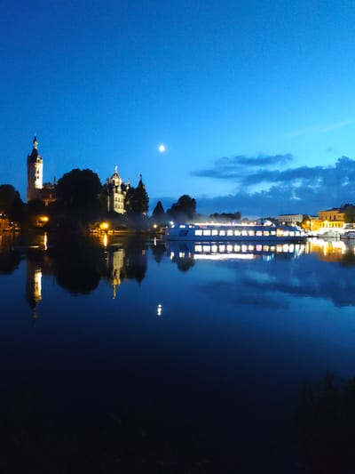 This image depicts a nighttime scene of the Schwerin Castle and its reflection in the waters of the Schweriner See in Schwerin, Germany. The sky is a deep blue, transitioning to a lighter blue near the horizon, with a few wispy clouds and a full moon visible. The castle, brightly illuminated, stands prominently on the left side of the frame, its towers and spires detailed against the dark sky. Its reflection is sharp and clear in the calm water below. A large passenger boat, also illuminated with many lit windows, sails across the water in the middle ground, its lights reflecting on the surface. On the right side of the image, more buildings are visible along the shoreline, also illuminated and reflecting in the water. The overall atmosphere is serene and picturesque, characteristic of a twilight or early night view of a historic waterfront. There are no people clearly visible in the image.