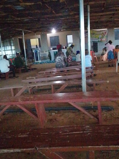 The image depicts an indoor gathering, likely a church service, in Rikina, Nigeria. The space appears to be a large hall with a rustic, unfinished ceiling constructed of corrugated metal and exposed wooden beams. The walls are painted white, and there are multiple windows with barred panes. The flooring is dirt.
In the foreground, several rows of wooden picnic-style benches are set up. Some are painted red, others appear to be unfinished wood. The benches are occupied by various individuals. A man in a white shirt is seated on a bench to the left. Further back, several people are seated, including a man in a dark camouflage-patterned shirt and a young person wearing a white t-shirt with blue lettering. A banner with colorful graphics and text, possibly related to a church event, is visible on the back wall.
In the mid-ground, a white podium with a blue trim stands near a table covered with a white cloth, suggesting an altar area. Candles are lit on the table. Several individuals are gathered around this area, some appearing to be participating in a service. To the right, more people are seated, and a small child in a pink shirt and blue skirt is standing.
The lighting is somewhat dim, with artificial lights illuminating the space. The overall impression is that of a community gathering in a humble, functional setting. The environment is consistent with a developing area in Nigeria, where resources might influence the construction and furnishings of public spaces.