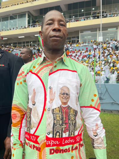 A man stands in a crowded stadium, likely in Malabo, Equatorial Guinea. He wears a custom-made button-down shirt featuring an image of Pope Leo XIV and the words "Papa León XIV Denos La Paz." The stadium is filled with people, many wearing white and yellow. Another man in a black suit with a badge is partially visible to the right of the main subject.