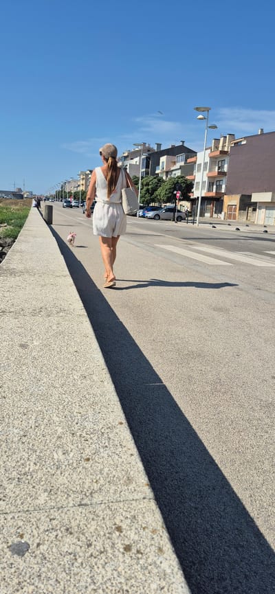 A woman, from behind, is walking along a paved promenade in Vila do Conde, Portugal, on a sunny day. She is wearing a light-colored romper, a cap, and sandals. She carries a large tote bag over her shoulder. A small dog, wearing a pink accessory, walks a short distance in front of her on a leash. The promenade runs parallel to a road with parked cars and some pedestrian traffic. Across the road, buildings with balconies and windows are visible, suggesting an urban or semi-urban coastal setting. The sky is clear and blue. The sun casts a long shadow of the woman and her dog across the promenade.