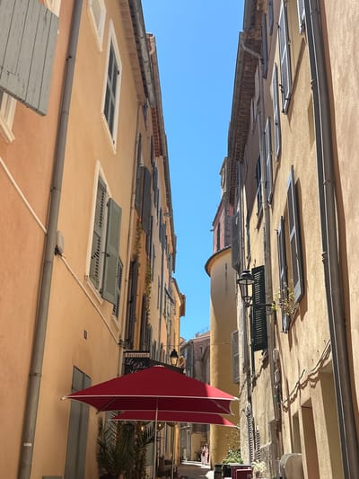 The image depicts a narrow, sun-drenched street in Saint-Tropez, France. The buildings lining the street are tall and painted in warm, earthy tones of yellow and orange. Many of the windows are adorned with closed, louvered shutters in shades of blue and gray, characteristic of Mediterranean architecture. A bright red umbrella, partially visible in the foreground, shades an outdoor seating area for a restaurant. A sign for "Restaurant Le Peche" is hanging on the wall to the left. The street itself is paved and slopes gently upwards, disappearing into the distance. The sky above is a vibrant, cloudless blue. The overall impression is one of a classic, charming European street scene bathed in strong sunlight.