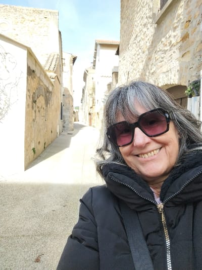 A smiling woman is captured in a selfie, positioned in a narrow, sunlit alleyway within what appears to be an old European town. She is a mature individual with gray hair, worn loose with bangs, and is wearing large, stylish black-framed sunglasses with a subtle reddish tint to the lenses. Her expression is joyful, with a wide smile that reveals her teeth. She is dressed in a black, padded winter jacket with a visible textured, possibly sherpa, lining around the collar and along the zipper. A black strap, likely from a bag, is visible across her shoulder.
The background shows a quaint street lined with tall buildings made of light-colored, rough-hewn stone or stucco, typical of historic architecture. The alley recedes into the distance, creating a sense of depth. Sunlight streams down, casting clear shadows on the stone-paved ground of the street, indicating a bright, clear day, likely late morning or early afternoon. On the left side, the building wall features faint, light-colored outlines or faded drawings. Further down the alley, small patches of greenery or moss cling to the lower parts of the walls. The sky visible above the buildings is a pale, clear blue. The overall impression is one of a pleasant, sunny day spent exploring a picturesque, historic location.