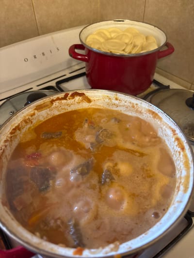 The image shows two pots on a white Orbis brand gas stovetop, likely in a kitchen. The stovetop is dirty. The kitchen is in Villa General Belgrano, Argentina. A red pot on the left contains uncooked dumplings. A larger pot to the right contains a stew or soup with visible pieces of meat, vegetables, and broth. The stew is simmering.