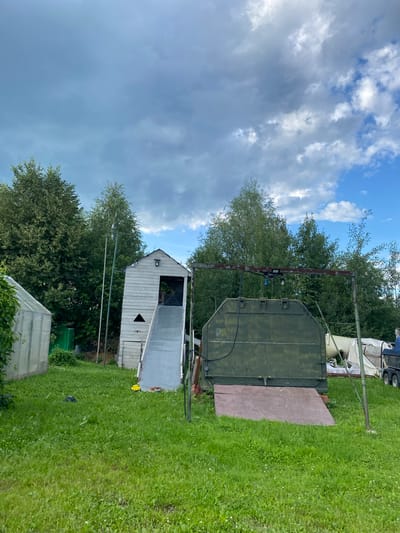The image depicts a backyard scene with various structures and natural elements. In the foreground, a lush green lawn is visible, with patches of white clover. To the left, a portion of a greenhouse with a translucent roof and white frame is present. Behind the greenhouse, a dark green fence can be seen.
Dominating the center of the image is a tall, slender wooden structure resembling a playhouse or observation tower. It is painted white and has a dark opening at the top. A long, grey slide extends from the opening down to the ground. Next to this structure, two thin, vertical poles stand close together, with what appears to be rope hanging between them.
To the right of the wooden structure is a large, dark green, irregularly shaped object, possibly a repurposed industrial container or part of a larger machine. It has visible hinges and latches. A wooden ramp leads up to its front. Behind this green object, there are white tarpaulins and other indistinct structures. In the far right of the image, a portion of a trailer with wheels is visible.
The background is filled with a dense line of mature green trees. The sky above is a mix of blue sky and heavy, grey clouds, suggesting potentially changeable weather.
The image does not contain any people or any explicit indication of the city of Novyi Ural, Russia. The scene appears to be a private outdoor space, possibly a backyard or a workshop area.