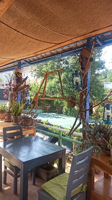 An outdoor dining area features a gray table and chairs, one chair with a green cushion. Above, a rustic wooden bicycle decoration hangs amidst lanterns and tree branches. A woven sunshade covers part of the area. The background shows lush green foliage and glimpses of buildings, suggesting a garden or patio setting.