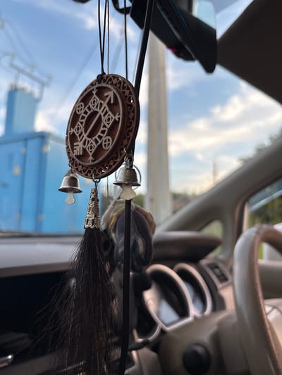 The image shows a wooden car charm hanging from the rearview mirror of a vehicle. The charm is circular with intricate geometric carvings and two small silver bells attached. A dark tassel hangs below the charm. The interior of the car is visible, including the dashboard, steering wheel, and a portion of the passenger seat. Outside the car, a bright blue industrial building is visible against a sky with white clouds. The scene appears to be an outdoor setting, possibly a parking lot or roadside. The location of Chigiri, Russia, is not discernible from the image.