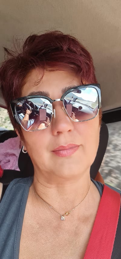A close-up selfie shows a woman with short, auburn hair wearing large, dark-rimmed sunglasses and a light pink lipstick. She is in the driver's seat of a car, as reflected in her sunglasses, with her hands on the steering wheel and her phone in view. A gold necklace with a small, clear pendant rests on her décolletage. She is wearing a grey top and a red seatbelt is visible across her chest. The background is blurred, suggesting movement. The location is not specified in the image.