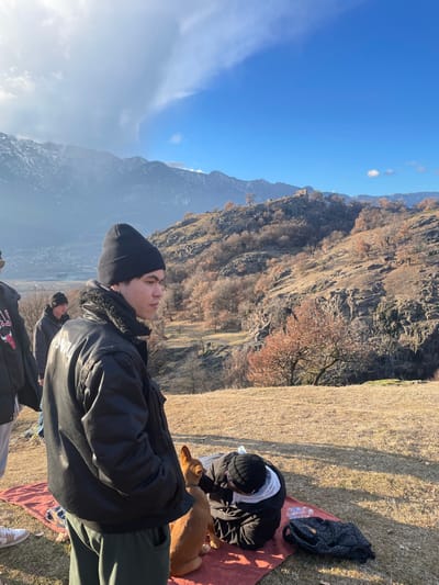 This image captures an outdoor scene set in a mountainous, natural environment under a clear, bright sky. The perspective suggests a landscape view, though the image is rotated 90 degrees counter-clockwise.
In the foreground, on the left side of the correctly oriented image, a young man is standing, looking towards the right. He has dark hair and is wearing a black beanie, a black jacket with a fur-lined collar, and dark pants. His face is illuminated by bright sunlight, suggesting he is looking towards the sun or a brightly lit distant view.
On the right side of the foreground, another person is seated on a reddish-orange blanket spread out on dry, yellowish-brown grass. This person is seen from behind, also wearing a black beanie and a dark jacket, and appears to be leaning forward, interacting with a light brown dog that is lying on the blanket or close beside them. A black bag, possibly a tote or backpack, and a clear plastic water bottle are visible near the blanket.
The midground is dominated by a steep, rocky mountainside covered with sparse vegetation. Many of the deciduous trees are bare or have dry, reddish-brown leaves, indicating late autumn or early winter. The rocky cliffs show varying textures and shades of brown and grey. A faint, distant structure, possibly an old building or ruin, can be seen on a peak further back.
In the background, beyond the immediate mountainside, a range of larger, more distant mountains stretches across the horizon. These distant peaks appear somewhat hazy, with patches that might be snow or cloud cover. The sky above is a vibrant blue with a few scattered white clouds, particularly towards the left.
The overall impression is one of a serene and sunny day spent in nature, possibly during a hike or a leisurely stop to enjoy the mountain views. The strong sunlight casts clear shadows and highlights the textures of the landscape and the individuals. No discernible text is visible on clothing or objects.