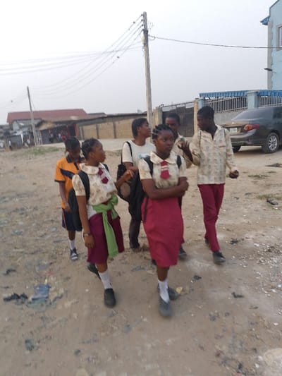 The image depicts a group of six young individuals, likely students, walking along an unpaved dirt road under an overcast sky, suggesting either early morning or late afternoon. The scene appears to be set in a semi-urban or residential area in Ijaiye, Nigeria, characterized by local architecture and infrastructure.
In the foreground and midground, three girls are prominently featured. Two are dressed in school uniforms, consisting of light-colored patterned short-sleeved shirts with red ties and dark red skirts or shorts. One of these girls has a bright green cloth tied around her waist. The third girl wears a white t-shirt and carries a black backpack. Accompanying them are three boys. One boy is seen in a yellow and blue striped t-shirt and dark shorts. Another wears a light-colored, patterned long-sleeved shirt with red trousers. A third boy is partially visible behind the girl in the white t-shirt. The group appears to be walking together, possibly to or from school, with some individuals seemingly engaged in conversation.
The immediate surroundings consist of the rough, unpaved road with scattered debris. In the background, various buildings are visible, including a light blue structure on the right side and several other buildings with light brown roofs. A dark-colored sedan car is parked next to a fenced area. Overhead, a utility pole with numerous power lines extends across the frame, indicating typical local infrastructure. There is no legible text visible on any signs or objects within the image. The overall mood is casual, with the individuals focused on their movement and interaction.