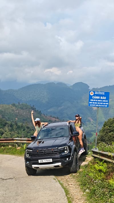 Two women in swimwear stand on and around a black Ford truck parked on a narrow road in a mountainous region. The truck has a license plate that reads "29K-179.83". One woman is standing on the truck's roof, gesturing with her arm raised. The other woman is standing on the truck's step, near the open door. Behind them, a blue road sign with white text in Vietnamese is visible. The sign reads "CẢNH BÁO", which translates to "Warning". The background shows a lush, green, mountainous landscape under a cloudy sky. This image was taken near Suối Giàng, Vietnam.