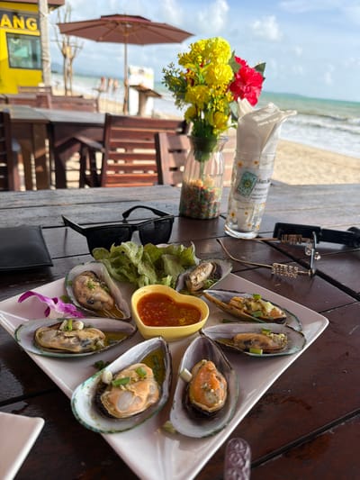 A plate of cooked mussels, a small bowl of sauce, a small bunch of salad, sunglasses, and a purple flower are arranged on a wooden table. In the background, a beach umbrella, chairs, a building, and the ocean are visible. The scene is set in Bang Muang, Thailand.
