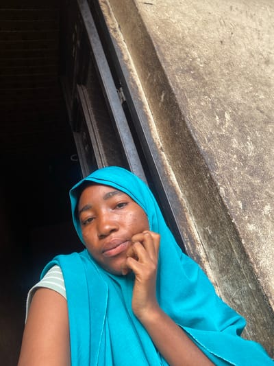 A young woman wearing a turquoise hijab and a striped shirt is seen from the chest up, looking towards the camera. Her left hand is resting on her chin. She is positioned in front of a dark, textured wall with a metal frame running vertically. The overall scene appears to be outdoors, possibly in a doorway or alcove. The image quality is slightly grainy, with a shallow depth of field that blurs the background.