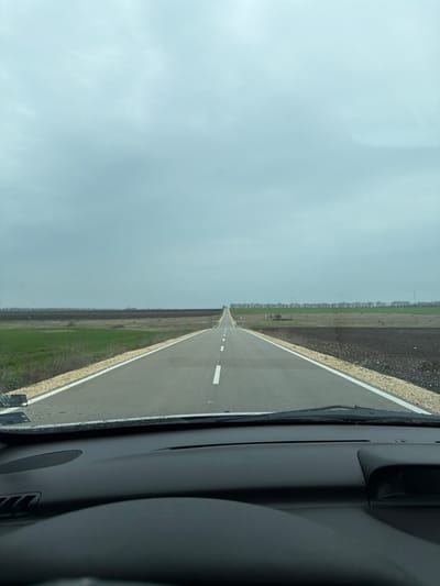 The image depicts a straight asphalt road extending into the distance under an overcast sky, viewed from the interior of a vehicle. The location is Gorun, Bulgaria. The road features a dashed white center line and solid white shoulder lines, flanked by light-colored gravel shoulders. To the left, a strip of green vegetation borders the road, followed by a field of dark brown, tilled earth. A distant line of trees forms the horizon on the left. To the right, a field of dark brown, tilled earth extends from the road, with a strip of green vegetation and a line of trees visible further back. A wind turbine is faintly discernible in the far distance on the right. The sky is uniformly light grey and cloudy. Water droplets are present on the vehicle's windshield, particularly visible along the lower edge. The vehicle's black dashboard, including vent grilles and a speaker grille, and the top portion of a steering wheel are visible in the foreground. No individuals or other vehicles are present within the visible scene.