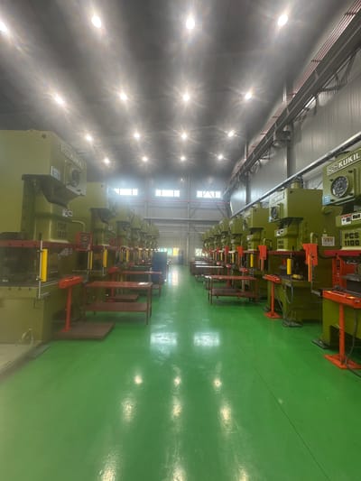 The image shows a row of industrial press machines in a factory setting. The machines are large, green, and appear to be of the "KUKIL" brand, with some labeled "PCS 1600" and "PCS 1100". They are arranged in a line, with their operational fronts facing the viewer. Red safety guards and metal shelving units are visible around some of the machines. The floor is a bright, glossy green. The ceiling is high with rows of circular fluorescent lights, some of which are creating glare. Windows are visible in the far wall. The overall environment is clean and organized, suggesting a manufacturing or production facility. The scene is located in the city of Pyeongtaek-si, South Korea, within a factory. There are no people visible in the image.