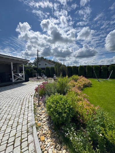 The image shows a sunny day in the city of Oliki, Russia, with a well-maintained backyard. A stone pathway leads to a covered patio area on the left. The backyard features a neatly mowed lawn, a variety of flowering bushes, and a hedge lining the back fence. In the distance, a two-story house is visible. There are no people visible in the image. The sky is blue with fluffy white clouds, indicating fair weather.