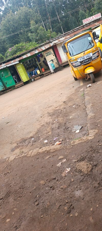 A yellow auto rickshaw with license plate KTWD 440J sits on a dirt road next to a row of small shops. The shops have corrugated metal roofs and green, yellow, and red accents. One shop has the name "TRAVIS KINYOZI" painted on the side. Another has a sign reading "DUKA LA VIOO." Shoes and other items are displayed outside the shops. Tall trees are visible in the background. The road is muddy and has tire tracks.