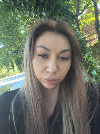 The image is a close-up selfie of a woman with light brown hair with blonde highlights, wearing a black shirt. She is outdoors, with lush green foliage and trees in the background. Part of a rustic wooden structure with a corrugated metal roof is visible behind the trees on the right side of the frame. A road and what appears to be a body of water or reflective surface are visible in the lower left quadrant. The lighting suggests it is daytime. The woman's face is angled slightly downwards, and she is looking towards the camera. Her makeup is visible, including eyeliner and mascara. The location is identified as Abzhanda, Abkhazia.