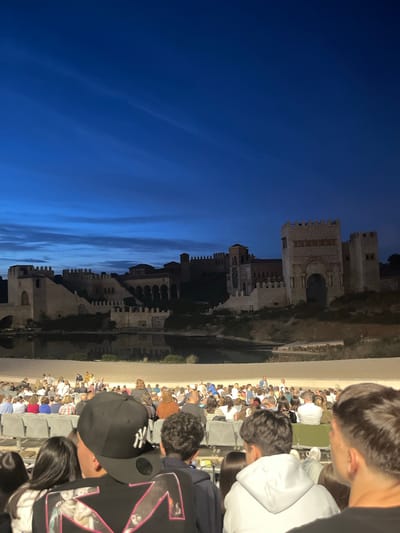 The image depicts a crowd of people seated in stadium-style seating, possibly at an event or performance in Toledo, Spain. To the left of the crowd, there is an elevated view of the city's historical architecture, particularly a well-lit medieval castle or fortress complex with multiple towers, crenellations, and arched structures. The sky above the fortress is a deep blue, suggesting either dusk or night, with some cloud streaking. The people in the crowd are generally facing towards the castle, and their attire indicates a casual setting. Several individuals are prominently visible in the foreground. The environment shows the transition from a man-made structure to natural sky.