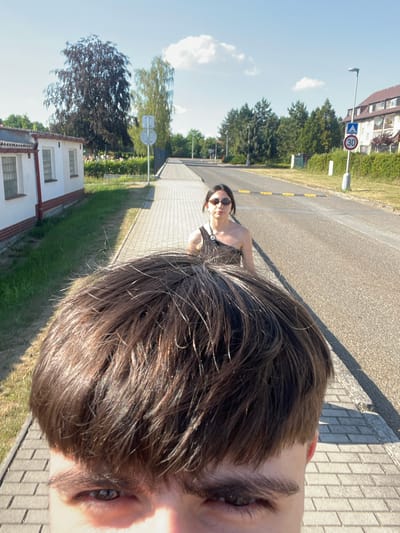 The media file depicts a close-up shot of a person's head and hair in the foreground, with a woman in the background. The scene appears to be outdoors, possibly in a suburban or rural area, with a street and sidewalk visible. The time of day seems to be daytime, with clear blue skies and bright sunlight casting shadows.

In the foreground, the focus is on the hair, which is dark brown and appears somewhat tousled. The person's eye and part of their face are also visible, suggesting the photo is a selfie taken from a low angle. The woman in the background is wearing sunglasses and a dark top. She is standing on the sidewalk, looking towards the camera.

The background shows a paved road, a sidewalk with brick paving, and a grassy area with trees. There are buildings visible in the distance, including a white building with red trim and windows, and a taller building that might be an apartment complex. A street light and a speed limit sign indicating "30" are also present. The overall impression is of a casual outdoor setting, possibly a walk or a moment captured while traveling. The mood seems relaxed and informal.

There is a visible speed limit sign with the number "30" inside a red circle and a blue arrow pointing upwards. The location context provided is Chotěšov, Czechia.
