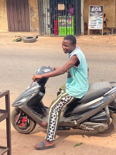 An outdoor scene in Neni, Nigeria, depicts a young adult male seated on a dark gray scooter, positioned slightly off the asphalt road on a dirt shoulder. The male, of African descent, wears a light blue sleeveless top with striped trim, olive green track pants featuring a distinct white and black geometric pattern down the sides, and dark slides. He is smiling and looking towards the camera, with both hands on the scooter's handlebars. The scooter displays a visible Kymco logo on its side and has a red front brake caliper.
In the background to the left, a light brown building with a large closed brown metal rolling door is visible. A discarded tire lies on the ground in front of it, along with some sparse vegetation. Further right, behind the male subject, a storefront is secured by a dark metal security gate. Inside, two individuals are partially visible: one in a pink top and another in red clothing, near a bright green display unit. A small, handwritten sign hangs from the gate.
To the far right, a white A-frame sign advertises "EZENWATA POS AVAILABLE" and "CHARGE YOUR PHONE & OTHER DEVICES HERE," detailing services such as money transactions and bill payment. A small child is partially visible near the base of this sign, adjacent to a wooden bench. The overall scene indicates a commercial street environment under daylight conditions.