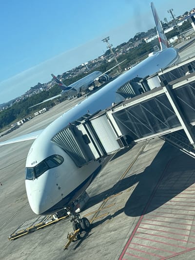 The image displays an airport tarmac scene, angled approximately 15-20 degrees counter-clockwise from horizontal. In the foreground, a white British Airways wide-body passenger aircraft is positioned, identifiable by the distinctive red, white, and blue livery on its tail fin and partially visible text "WBA" on the cockpit frame, and "TEM PROTAIR" on the lower fuselage. A flexible passenger jet bridge is extended and connected to the forward door of this aircraft. A yellow tow bar is attached to the nose landing gear, and a ground support equipment conveyor belt is positioned near the front cargo hold. The tarmac is concrete/asphalt, marked with yellow lines and red striped areas. In the middle ground, another white passenger aircraft, identifiable as a LATAM airline plane by the branding on its engine and tail fin, is visible, appearing further back on the tarmac. The background features a clear blue sky over a hilly landscape densely populated with residential structures and interspersed with green vegetation. Airport light poles or towers are visible across the scene. The lighting indicates strong daylight, casting pronounced shadows on the tarmac. The urban and airport context is consistent with the city of Guarulhos, Brazil. No individuals are discernible in the frame.