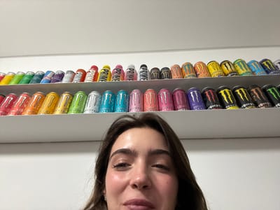 A woman is upside down, with her face appearing at the top of the frame. Below her are two rows of energy drinks on a shelf. The top row features primarily Monster energy drinks in various colors. The bottom row includes a mix of brands, with Prime visible. The background appears to be a white wall.
