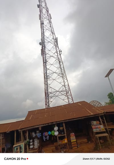 A telecommunications tower looms over a cluster of rustic buildings in Nguroje, Nigeria. The tower, constructed of metal lattice, extends high into a cloudy sky, dotted with several antennas. Beneath the tower, corrugated metal roofs, weathered to a rusty brown, cover a collection of shops. These open-fronted establishments display various goods, including buckets, cookware, and bottled beverages, suggesting a bustling marketplace. A solar-powered street lamp is visible to the right of the tower.