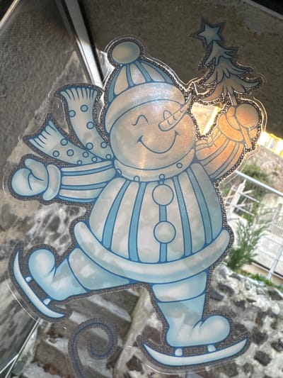 A transparent window cling, depicting a light blue and white snowman with a glitter outline, is affixed to a clear glass pane. The snowman, presented in a dynamic pose, wears a striped winter hat and a polka-dotted scarf. Its face features a carrot nose, two dots for eyes, and a wide, upturned smile. Its torso is vertically striped with three circular buttons. The snowman's raised right arm holds a stylized Christmas tree topped with a star, while its left arm is bent. It wears ice skates, with a spiral trail visible beneath the left skate, indicating movement. Through the window, an out-of-focus exterior scene reveals a textured grey surface to the upper left and center, and green foliage with a metal railing to the right. The scene is illuminated by natural light and is located in the city of Le Puy-en-Velay, France.