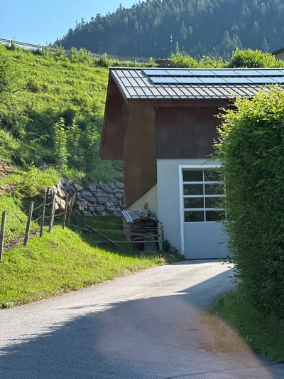 This image captures a view of a modern building with solar panels on its roof, situated on a grassy, tree-covered hillside in Annaberg-Lungötz, Austria. A paved road leads towards the building, with a fence line along the left side of the road, bordering a steep, grassy incline. The building features a dark brown wooden facade and a light-colored section with a large garage door and a window with multiple panes. A dense, green hedge is visible on the right side of the image. The sun casts a long shadow across the road. The overall scene is one of a rural residential or agricultural setting bathed in bright sunlight.