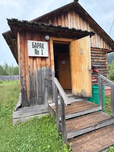 A weathered wooden building, identified by a sign as "БАРАК № 1" (Barak No. 1), stands prominently. The building is constructed from logs and planks, with a dark, aged appearance. An open wooden door reveals a dark interior, suggesting it might be a dwelling or a utility structure. A wooden staircase leads up to the entrance, and a green barrel sits to the right of the stairs. The surrounding environment is characterized by lush green grass, clover, and some wildflowers, indicating a rural or natural setting. In the background, a wooden fence and trees are visible, hinting at the location being on the outskirts of a settlement. The sky is overcast with visible raindrops, suggesting recent or ongoing rain. The scene appears to be in the village of Ust'-Pyzas, Russia, given the context of the request, implying a remote, possibly rustic, and sparsely populated area.