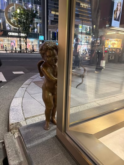 A bronze statue of Cupid, the Roman god of love, stands on a tiled sidewalk in front of a building with a large glass storefront. The statue is partially obscured by the building's frame, with its right side hidden from view. Cupid is depicted as a child with wings, holding a bow and arrow. The background shows a street scene at night with neon lights and buildings. The location is identified as Chuo, Japan. People are visible in the background, walking on the street and reflected in the store window. A large tree grows next to the building, and a sign with the Giorgio Armani logo is visible on the storefront. The overall impression is that of a commercial street in a Japanese city at night.