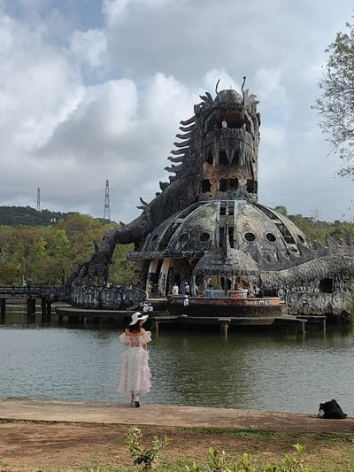 The image depicts the abandoned Thuy Tien Lake Water Park, located in Kim Long Ward, Hue City, Vietnam. A large, weathered, concrete and stone dragon structure is the central subject, rising from a still, green-tinted lake.
The structure features a towering dragon head, appearing to be part of a multi-level domed building, with the dragon's body wrapping around its base and extending into the water. The dragon's features are intricate, showing scales, claws (one large claw visible near the dome), and an open mouth. The surfaces are discolored and show significant signs of weathering and decay, with exposed structural elements and broken openings visible on the dome and within the dragon's head.
In the foreground, a female subject stands with her back to the viewer, positioned on a paved embankment at the lake's edge, facing the dragon structure. She is wearing a light-colored, possibly floral-patterned, off-the-shoulder dress with ruffled sleeves and a wide-brimmed white hat. To her right, a dark bag rests on the ground.
Multiple individuals are discernible on and around the dragon structure. One person is clearly visible standing within the elevated opening of the dragon's mouth. Other indistinct figures are present on the platforms and lower levels of the dome. A dark, concrete bridge extends from the left bank of the lake towards the base of the dragon structure. The background reveals dense green foliage on a distant hillside, topped by two metal lattice transmission towers. The sky is overcast with scattered grey and white clouds.