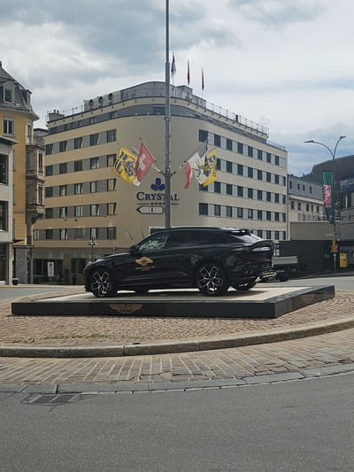 A black Aston Martin is displayed on a raised platform in front of a light-colored building. The building has multiple floors with rows of windows and a curved facade. Several flags are visible, including the Swiss flag and what appears to be a yellow and blue crest flag, as well as a red flag. The Aston Martin has a gold emblem on its side that reads "BRITISH CLASSIC CAR". A black sign with the Aston Martin logo is also visible next to the car. The ground in front of the building is paved with bricks, and a paved road runs alongside it. The sky is cloudy. The scene appears to be an outdoor display in a town or city, likely St. Moritz, Switzerland, given the contextual information. The time of day appears to be daytime, with bright lighting. No people are clearly visible in the frame.