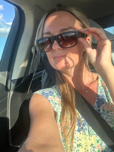 A blonde woman wearing sunglasses and a floral print dress is sitting in the passenger seat of a car. She is adjusting her sunglasses with her right hand. Her hair is long and blonde, and it is partially obscuring her face. The car appears to be in motion, as indicated by the blurred background. The sky is blue with some clouds. The woman is wearing a seatbelt. The image appears to have been taken during the daytime. The location is Novotolucheyevo, Russia.