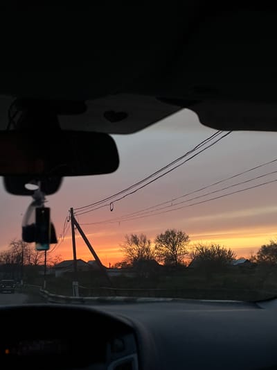 This image captures a view from inside a vehicle, looking out at a sunrise or sunset over a landscape in Alexandrovka, Russia. The sky is a prominent feature, displaying a vibrant gradient of colors, from bright orange and yellow near the horizon on the right, transitioning to soft pinks and grays across the rest of the sky. Long, horizontal cloud streaks are visible in the upper sky, catching the light.
In the midground, several bare trees are silhouetted against the colorful sky, suggesting late autumn, winter, or early spring. Utility poles with multiple power lines stretch across the scene, adding an element of human infrastructure to the natural beauty. Faint outlines of distant buildings, possibly houses, can be seen nestled among the trees, indicating a rural or semi-rural setting.
The foreground is dominated by the dark interior of the car, including parts of the dashboard and the silhouette of a rearview mirror on the left. A smaller device, possibly a dashcam or phone mount, is also silhouetted towards the bottom-left. The overall atmosphere conveys a sense of peaceful observation during the transitional period of dawn or dusk. No visible text is discernible in the image.