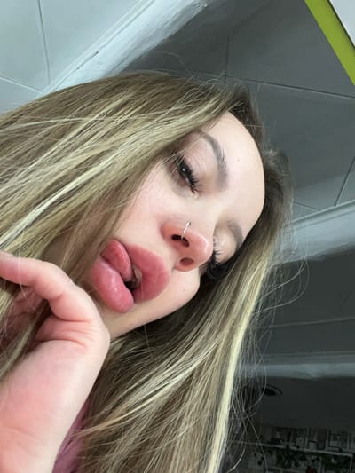A close-up, tilted photograph depicts a young woman with long, blonde hair and a silver nose ring. Her lips are slightly parted with her tongue partially visible. Her left hand is raised near her face, with only her fingers visible. The background shows light gray walls with white trim. The specific location in Port-Katon, Russia is not discernible from the provided image.