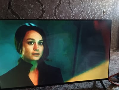 The image shows a television screen displaying a scene with a woman in the foreground. She has dark hair and is looking to the left of the frame. The background on the screen is dark and abstract, with a bright yellow and red-orange area on the right side. The television is placed in a room with patterned wallpaper and a carpeted floor. There is also a small electrical outlet visible on the wallpaper. The image does not provide any information about the location in Bogorodsk, Russia.