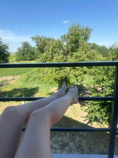 This media file features a close-up view of a person's legs and feet resting on a black metal railing. The person's skin is fair, and their feet are bare. The background consists of lush green trees and a clear blue sky, suggesting a daytime outdoor setting. The scene is framed by the railing, with shadows of leaves cast upon the ground behind it. The overall impression is one of relaxation and enjoying a warm, sunny day. The location is identified as Yablunivka, Ukraine. There is no visible text in the media file.