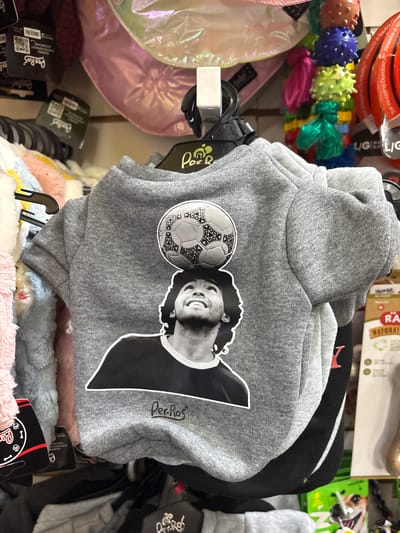 The image shows a gray hoodie for dogs hanging on a rack in a store, likely in La Plata, Argentina, given the hoodie's design. The hoodie features a black-and-white image of Diego Maradona next to a soccer ball, suggesting a connection to Argentina's football culture. The hoodie is labeled "Per-Ros," possibly a brand name. Other items are visible on the rack, including dog toys and other pet supplies. The scene depicts a retail environment specializing in pet products.