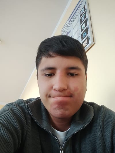 A young male individual, centrally positioned, is depicted in an indoor setting within Rishtan, Uzbekistan. He has short, dark hair and gazes downwards and slightly to his left, exhibiting a facial expression with pursed lips. Reddish skin patches are visible on both cheeks and the chin. He wears a dark grey, ribbed, zip-front jacket over a white undershirt. The background features light-colored walls and a white ceiling with a partial view of an overhead light fixture in the upper left. A rectangular information board, angled on the upper right wall, displays text in an unidentified script and several graphic icons, some resembling software application logos. The environment suggests an institutional or educational building.
