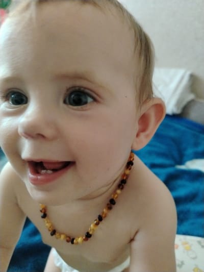 A close-up, waist-up shot shows a baby with fair skin and light brown hair. The baby is wearing a beaded necklace with amber and dark brown beads. The baby is looking to the left with a wide, open-mouthed smile, revealing small teeth. The background consists of a blurred blue blanket and a white sheet. The setting is an indoor environment, likely a home. The location in the city of Molodiia, Ukraine, is not discernible from the image.