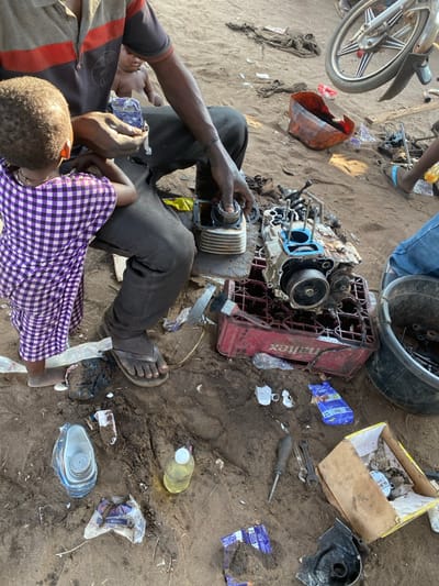 A mechanic, seated on sand, assembles an engine cylinder. A child in a purple checkered shirt stands nearby. Scattered around them are engine parts, tools, a red crate labeled "Haltex," boxes, and various plastic containers. A motorcycle wheel is partially visible in the upper left. The scene appears to be a repair shop, characterized by its dusty environment.
