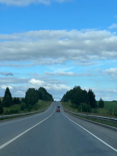This image depicts a rural highway landscape, likely located near Verkhne-Pozim', Russia, under a partly cloudy sky. The perspective is from a moving vehicle, looking forward along a two-lane asphalt road that curves gently to the right as it ascends a slight incline.
The road is marked with a solid white line on the right edge and a broken white line separating the lanes. Guardrails, constructed of metal, run along both sides of the road. On the left side of the road, a dense line of dark green trees, including coniferous and deciduous varieties, borders a grassy embankment. On the right side, the terrain slopes upwards, with more trees and patches of green fields visible.
Several vehicles are present on the road. In the foreground, a red car is visible in the right lane. Further ahead, in the distance, more cars can be seen, including what appears to be a black SUV in the left lane. Overhead, a highway sign structure with what looks like electronic messaging capabilities is positioned above the road in the distance. The sky is a bright blue, interspersed with scattered, fluffy white and grey clouds. The overall impression is one of a typical summer day on a highway in a wooded, rural area.