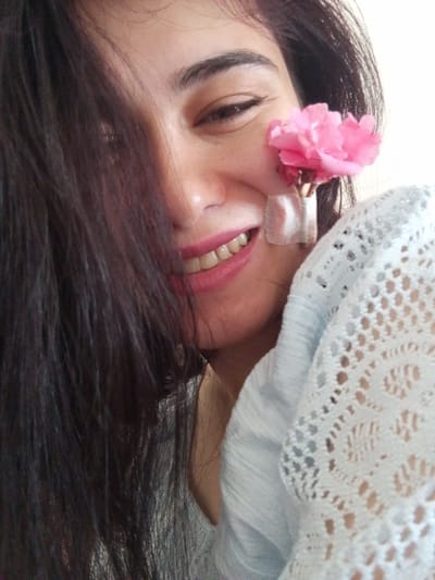 A close-up, selfie-style photograph shows a person's face adorned with a pink flower placed on their cheek, secured with two strips of medical tape. The person has dark hair that partially obscures their face and is smiling, revealing teeth. They are wearing a light-colored, lacy garment. The background is a plain, light-colored wall, suggesting an indoor setting. No specific landmarks or features of Be'er-Sheva are visible in the image.