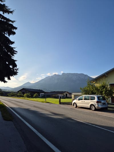 The image depicts a daytime scene in Niederndorf, Austria, featuring a road, a silver minivan, houses, green fields, and a mountain range in the background. A large conifer tree is visible in the foreground on the left. The road is paved and has a white line marking its edge. The minivan is parked on the right side of the road. Houses with yellow and brown facades are situated behind the minivan and across the road. The mountains in the distance are covered in vegetation and appear to be illuminated by the sun. The sky is clear and blue, with a few wispy clouds. The overall impression is one of a rural or semi-rural setting on a sunny day.