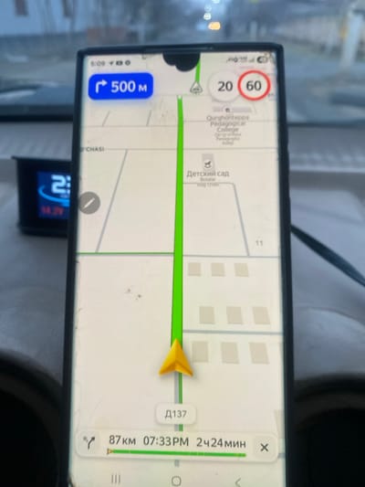 The image displays a dark-colored smartphone positioned on a beige vehicle dashboard, actively showing a navigation application. The screen is lightly smudged. The navigation map depicts a green route with a yellow directional arrow indicating the current vehicle position and trajectory. A blue notification banner in the upper left corner indicates "Turn right 500 M". Speed limit indicators of "20" (advisory) and "60" (regulatory, red-circled) are visible in the upper right corner.
Specific location details visible on the map identify the area within Kurgantepa (Qo'rg'onteppa), Uzbekistan, by labels for "Qurghonteppa Pedagogical College" (Qo'rg'onteppa Pedagogika Kolleji) and "Детский сад" (Kindergarten, also labeled Bolalar bog'chasi). A road segment is marked "D137". The navigation summary at the bottom displays "87 km" remaining distance, an estimated arrival time of "07:33 PM", and a remaining travel time of "2 ч 24 мин".
To the left of the smartphone, a smaller black electronic device is visible on the dashboard, displaying "2:.." and a voltage reading of "14.2V". The background, viewed through the vehicle's windshield, is blurred and shows an indistinct outdoor street environment with what appear to be structures or buildings.