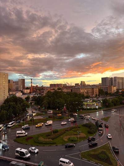 The image depicts a city intersection at dusk, with a dramatic sky filled with colorful clouds. The scene is a busy urban environment, with multiple lanes of traffic on wet asphalt roads, suggesting recent rain. Several cars, vans, and trucks are visible, navigating the roundabout and traffic lights. Green trees line the streets and a circular landscaped area with bushes and flowers is present in the center of the intersection. In the background, there are residential buildings, an industrial chimney, and other city structures. The lighting suggests the transition from day to night, with the sky illuminated by the setting sun. The location is identified as Tarasovka, Russia. There are no people clearly visible in the image.