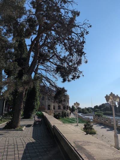 The image depicts a scene in Ganja City, Azerbaijan, specifically at the Nizami Park. In the foreground, a paved walkway, constructed with grey rectangular bricks, leads the eye towards a distant view. To the left, a large tree with dark, dense foliage dominates the frame, casting a prominent shadow across the walkway. A brown wooden bench is positioned under the tree, offering a place to rest. Beyond the walkway, a low stone wall with a railing separates the park from a body of water, likely the Ganja River, which is visible in the middle ground. A series of green chairs are neatly arranged on a grassy area beyond the wall, suggesting an outdoor seating or performance space. Several ornate lampposts, with multiple light fixtures, are visible along the walkway, contributing to the park's ambiance. In the background, a large, multi-story building with classical architectural elements stands prominently. The sky is a clear, bright blue, indicating a sunny day. The overall scene conveys a serene and picturesque park setting.