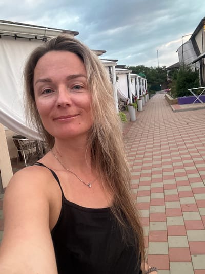 A woman with long blonde hair and a black tank top takes a selfie in front of an outdoor dining area in Yaroslavskaya, Russia. The woman is smiling and looking at the camera. The dining area has white tents with tables and chairs. The ground is paved with pink and white tiles. In the background, there are trees and a cloudy sky.